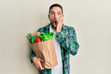 Wall Mural - Handsome man with beard holding paper bag with groceries afraid and shocked, surprise and amazed expression with hands on face