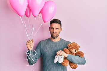 Poster - Handsome man with beard expecting a baby girl holding balloons, shoes and teddy bear clueless and confused expression. doubt concept.