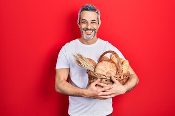 Wall Mural - Handsome middle age man with grey hair holding wicker basket with bread looking positive and happy standing and smiling with a confident smile showing teeth