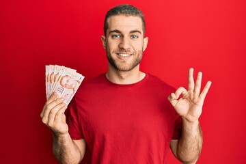 Poster - Young caucasian man holding 10 singapore dollars banknotes doing ok sign with fingers, smiling friendly gesturing excellent symbol