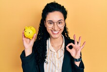 Young Hispanic Woman With Curly Hair Holding Piggy Bank Doing Ok Sign With Fingers, Smiling Friendly Gesturing Excellent Symbol