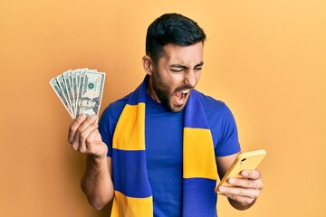 Poster - Young hispanic man football supporter using smartphone holding usa dollars banknotes angry and mad screaming frustrated and furious, shouting with anger. rage and aggressive concept.