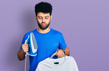 Sticker - Young arab man with beard holding electric steam iron and white t shirt making fish face with mouth and squinting eyes, crazy and comical.