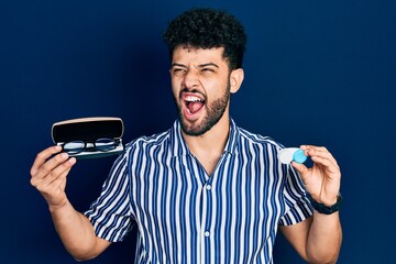 Canvas Print - Young arab man with beard holding glasses and contact lenses angry and mad screaming frustrated and furious, shouting with anger. rage and aggressive concept.