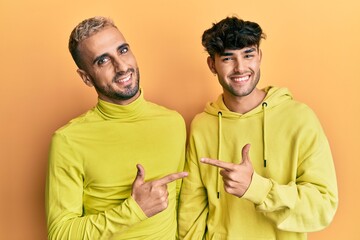 Poster - Homosexual gay couple standing together wearing yellow clothes cheerful with a smile of face pointing with hand and finger up to the side with happy and natural expression on face