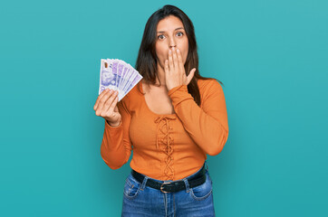 Poster - Beautiful hispanic woman holding 20 swedish krona banknotes covering mouth with hand, shocked and afraid for mistake. surprised expression