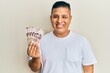 Young latin man holding 500 mexican pesos banknotes looking positive and happy standing and smiling with a confident smile showing teeth