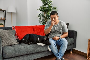 Wall Mural - Young latin man and dog sitting on the sofa at home feeling unwell and coughing as symptom for cold or bronchitis. health care concept.