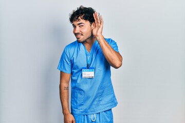 Sticker - Young hispanic man wearing blue male nurse uniform smiling with hand over ear listening an hearing to rumor or gossip. deafness concept.