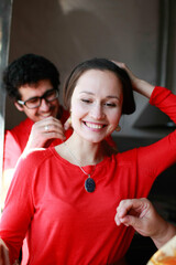 The man presented the woman with a necklace and puts the jewelry around her neck. The woman rejoices at the gift. Man and woman in red clothes. Mom supports young