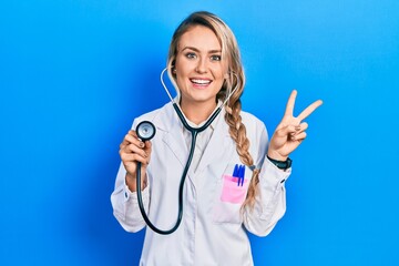 Sticker - Beautiful young blonde doctor woman holding stethoscope smiling looking to the camera showing fingers doing victory sign. number two.