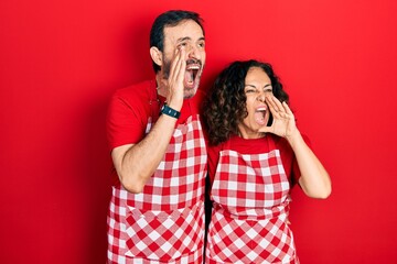 Wall Mural - Middle age couple of hispanic woman and man wearing cook apron shouting and screaming loud to side with hand on mouth. communication concept.