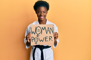 Poster - Young african american girl wearing karate kimono holding woman power banner looking positive and happy standing and smiling with a confident smile showing teeth