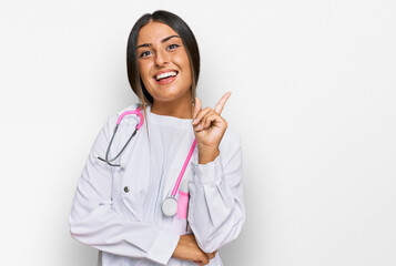 Wall Mural - Beautiful hispanic woman wearing doctor uniform and stethoscope with a big smile on face, pointing with hand and finger to the side looking at the camera.