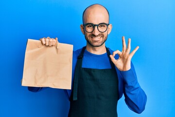 Young hispanic man wearing waiter uniform holding take away paper bag doing ok sign with fingers, smiling friendly gesturing excellent symbol