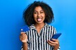 Beautiful african american woman with afro hair holding smartphone and credit card sticking tongue out happy with funny expression.