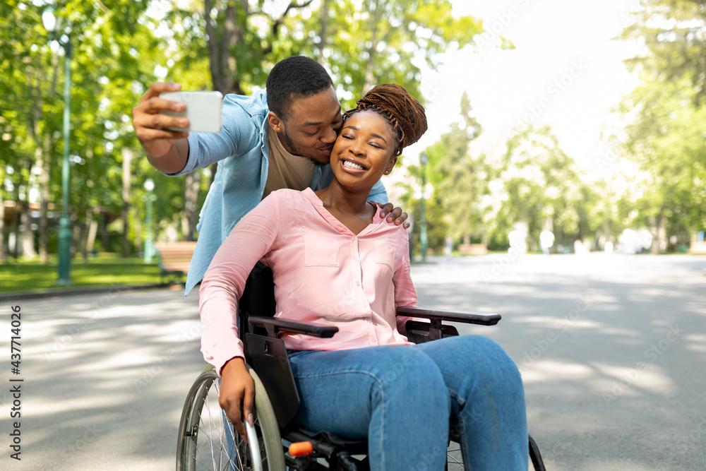 Romantic black man and his impaired girlfriend in wheelchair taking selfie together, kissing at city park - obrazy, fototapety, plakaty 
