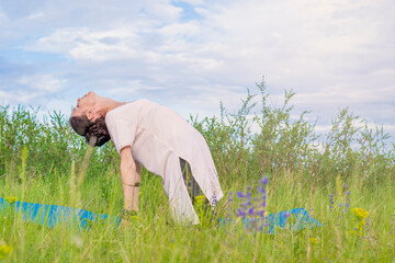 Wall Mural - A young woman sits on a green open-air lawn and performs backbend yoga exercises. Outdoor sports life.