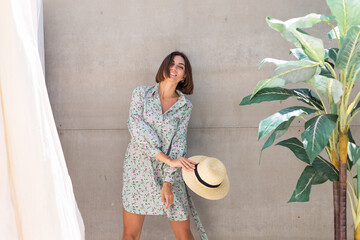 Wall Mural - Beautiful woman in summer dress and straw hat by palm tree and gray wall on background, playful joyful  positive excited cheerful happy