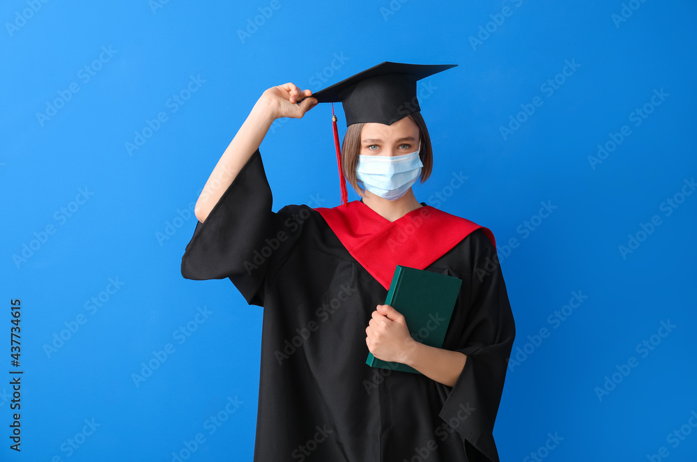 Female graduating student in protective mask and with book on color background