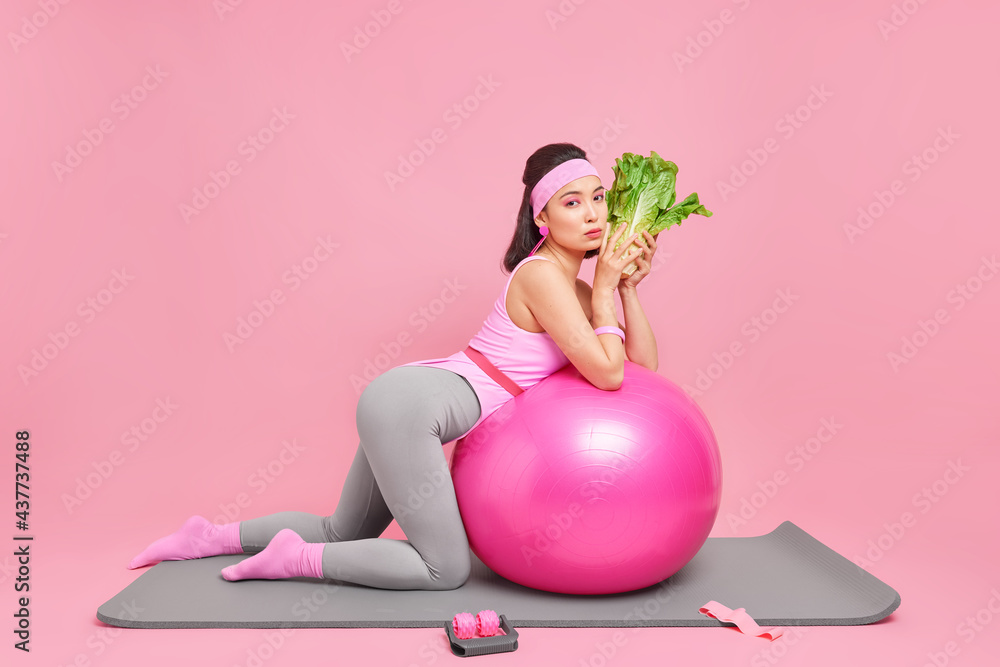 Sideways shot of serious motivated dark haired Asian sportswoman leans at swiss ball holds green vegetable eats healthy food leads active lifestyle has regular workout to be fit poses on fitness mat - obrazy, fototapety, plakaty 