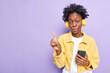 © WHstudio Leushin N - Studio shot of surprised woman with dark skin curly hair points at upper left corner uses smartphone application for listening music wears headphones on ears isolated over purple background.