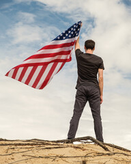 A man with an American flag stands on the sand, skies on the background. Man is holding waving American USA flag, left arm is up. Fourth of July Independence Day.