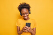 © WHstudio Leushin N - Studio shot of smiling teenage girl with dark skin sends text messages via smartphone chooses song from playlist listens music wears stereo headphones on ears isolated over yellow background
