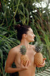 © verona_studio - Young beautiful girl posing on the tropical background, holding pineapples in hands.  Woman with bronze tan in bikini.