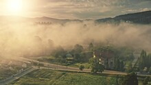 Sunny Morning In European Mountain Village. Drone Fly Through Yellow Mist, Sunrise Soft Light With Sun Beams In Background. Summer Nature Landscape. Cottages And Rural Road With Cars. Cinematic Shot