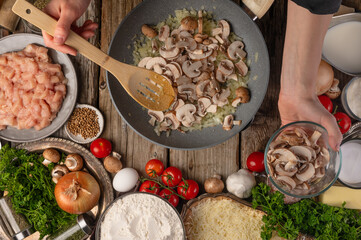 Wall Mural - The chef prepares pizza. In the photo, the process of cooking mushrooms. The cook mixes the mushrooms in a frying pan with a wooden spoon. There are many products on the table. View from above.