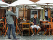 Young man standing taking a photo of his father who is sat at a chair in the outside of a beautiful and unidentifiable coffee shop. Dad being photographed by son outside in daylight.