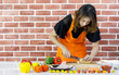 Beautiful young Asian woman in orange apron stand in front of brown brick wall of kitchen and carefully slicing hotdog on cooking table that full of fresh tomatoes, healthy bell peppers, and dish