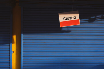 Hanging closed sign board in front of blue shutter door of grocery with sunlight and shadow on surface in vintage tone style