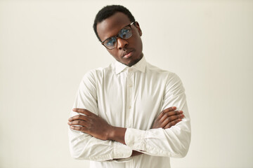 Horizontal shot of serious stubborn young black male in formal wear and eyeglasses keeping arms folded on his chest in closed posture, expressing disagreement or discontent. Body language