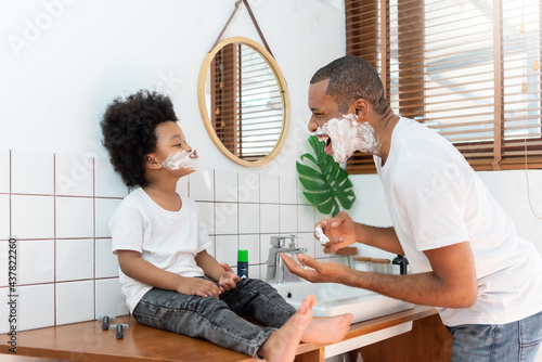 Funny Black African American Father and little boy laughing while playing shaving foam on their face in bathroom at home on holiday. Happy African family having fun