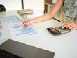 © phoomrat - Top view of hand pointing to data from business information charts and pressing calculator to calculate company's finances. Business woman analyzing financial report with calculator