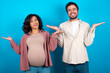 © Roquillo - Cheerful cheery optimistic young couple expecting a baby standing against blue background holding two palms copy space