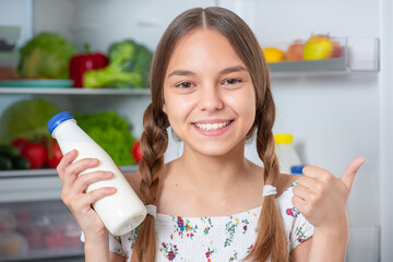 Wall Mural - Beautiful young teen girl holding bottle of milk and drinks while standing near open fridge in kitchen at home