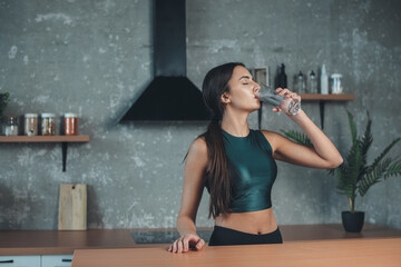 Caucasian woman wearing sportswear is drinking a glass of water to hydrate her body in the kitchen