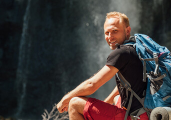 Wall Mural - sincerely smiling man portrait with backpack dressed in active trekking clothes sitting near mountai