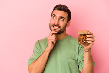 Wall Mural - Young caucasian man holding an almond jar isolated on pink background relaxed thinking about something looking at a copy space.