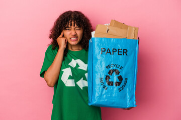 Wall Mural - Young caucasian woman holding a recycled plastic isolated on pink background covering ears with hands.