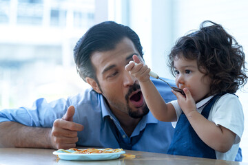 Adorable 2 years old girl eating spaghetti pasta enjoyment with their hands, and father sitting nearby