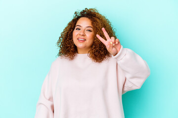 Wall Mural - Young latin curvy woman isolated on blue background joyful and carefree showing a peace symbol with fingers.