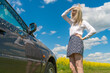 slender young blonde woman in a skirt and white blouse stands near a dirty car stuck on a dirt road in the middle of a rapeseed field, looks at the wheel and holds her head
