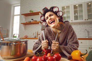 Funny crazy cheerful woman housewife in curlers and furry bathrobe standing in kitchen and holding kitchenware in hands during cooking. Funny women at home concept