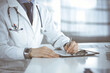 © rogerphoto - Unknown male doctor sitting and working with clipboard of medication history record in clinic at his working place, close-up. Young physician at work. Perfect medical service, medicine concept