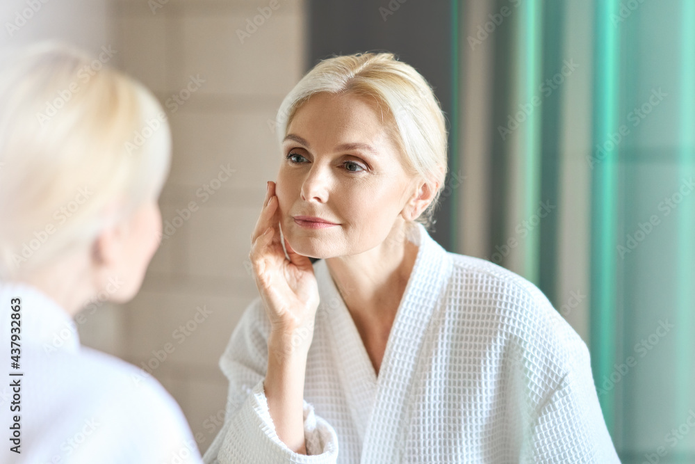 Gorgeous mid age adult 50 years old blonde woman standing in bathroom after shower touching face, looking at reflection in mirror doing morning beauty routine. Older dry skin care concept. - obrazy, fototapety, plakaty 