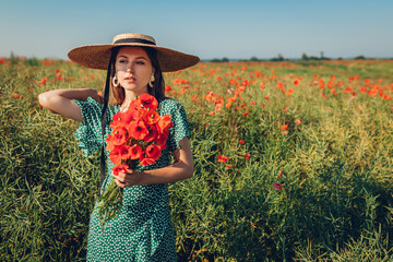 Wall Mural - Beautiful woman gathered bouquet of poppies flowers walking in summer field. Stylish girl wearing vintage straw hat